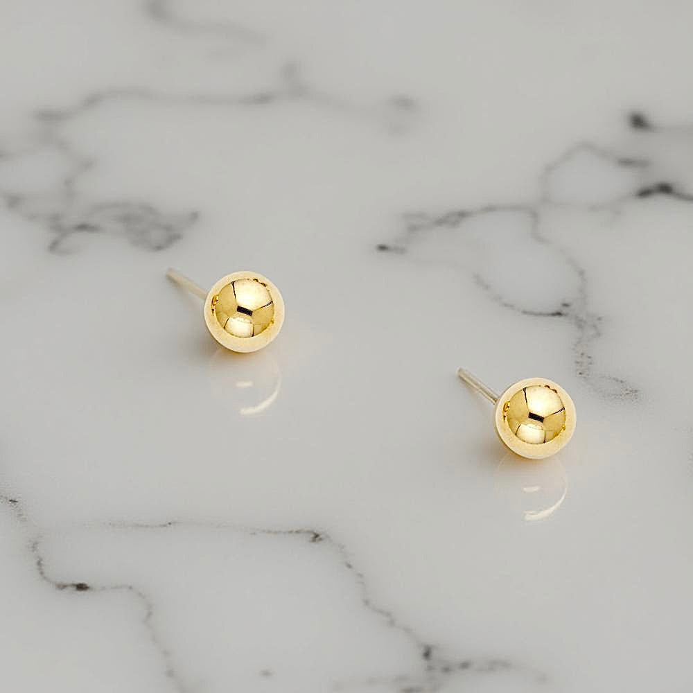 A close-up of 14K yellow gold 7mm ball stud earrings resting on a white marble background, highlighting their polished round surface.