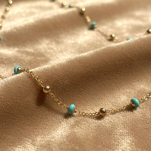 14K yellow gold necklace with turquoise enamel beads elegantly arranged on a fawn-colored velvet surface, showcasing the chain’s shine and colorful enamel accents.