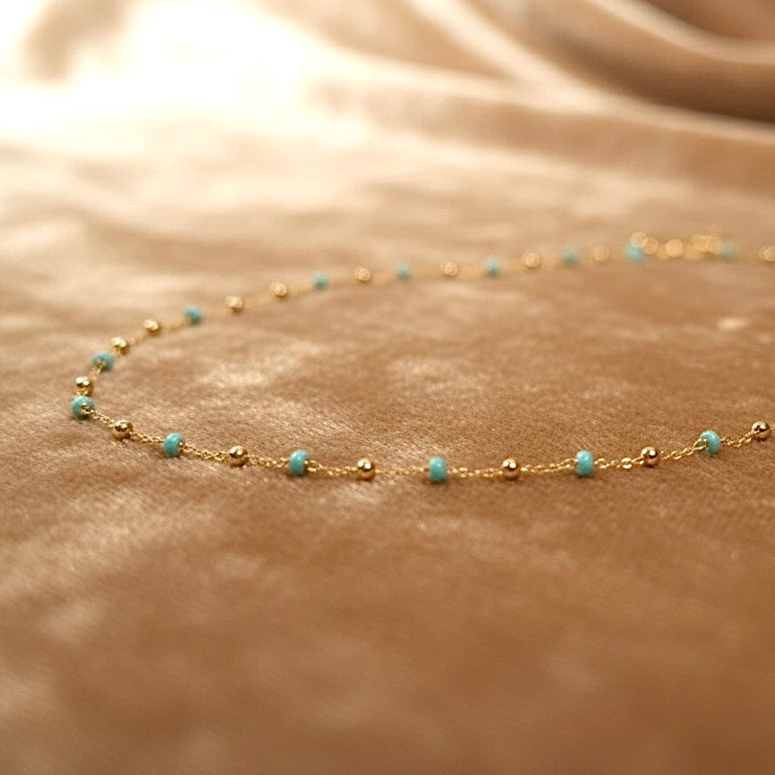 14K yellow gold necklace with turquoise enamel beads elegantly arranged on a fawn-colored velvet surface, showcasing the chain’s shine and colorful enamel accents.