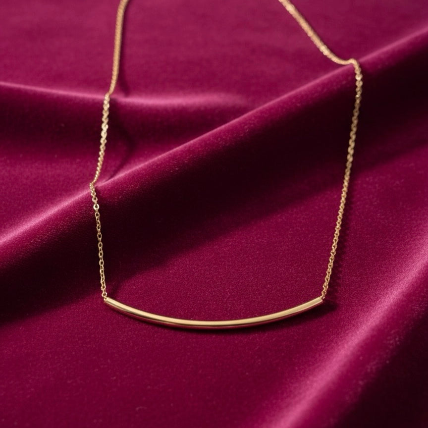 14K Yellow Gold Curved Bar Necklace laid on magenta velvet cloth, highlighting the smooth curve and warm gold shine.