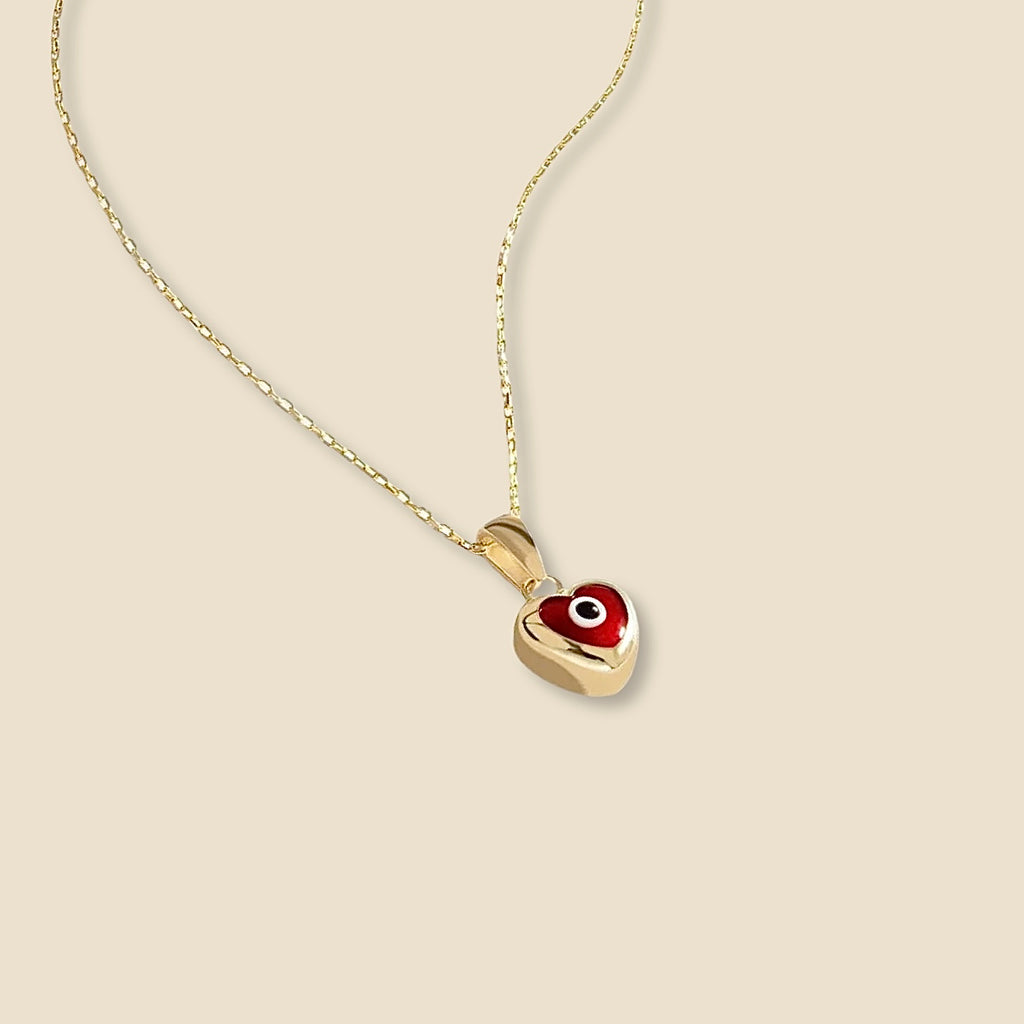 A close-up of the 14K yellow gold heart pendant with a deep red enamel eye center, resting on neutral and clean background, symbolizing classic protection and offering a minimalist yet bold pop of color.