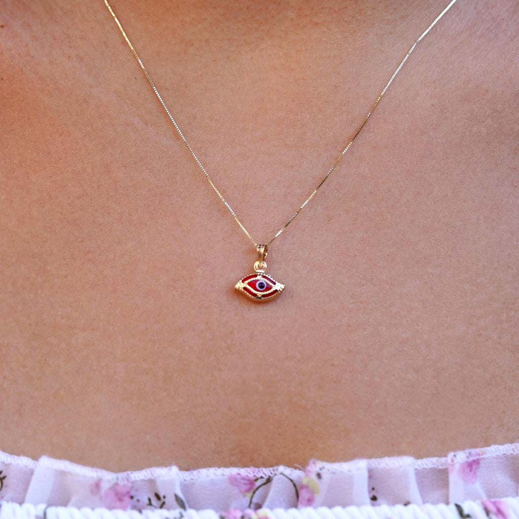 A model wearing a delicate 14K yellow gold red evil eye pendant featuring a brilliant red cubic zirconia center, resting elegantly on her neckline. The fine gold chain and polished finish reflect soft light, highlighting the pendant’s symbolic protection and timeless charm.