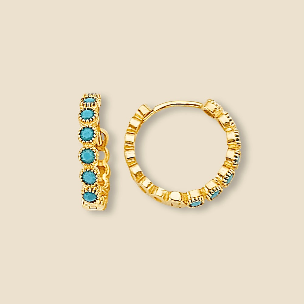Close-up of a pair of 14K yellow gold round turquoise Huggies earrings displayed on a plain background, showcasing polished gold hoops with vibrant turquoise stones set evenly along the surface.