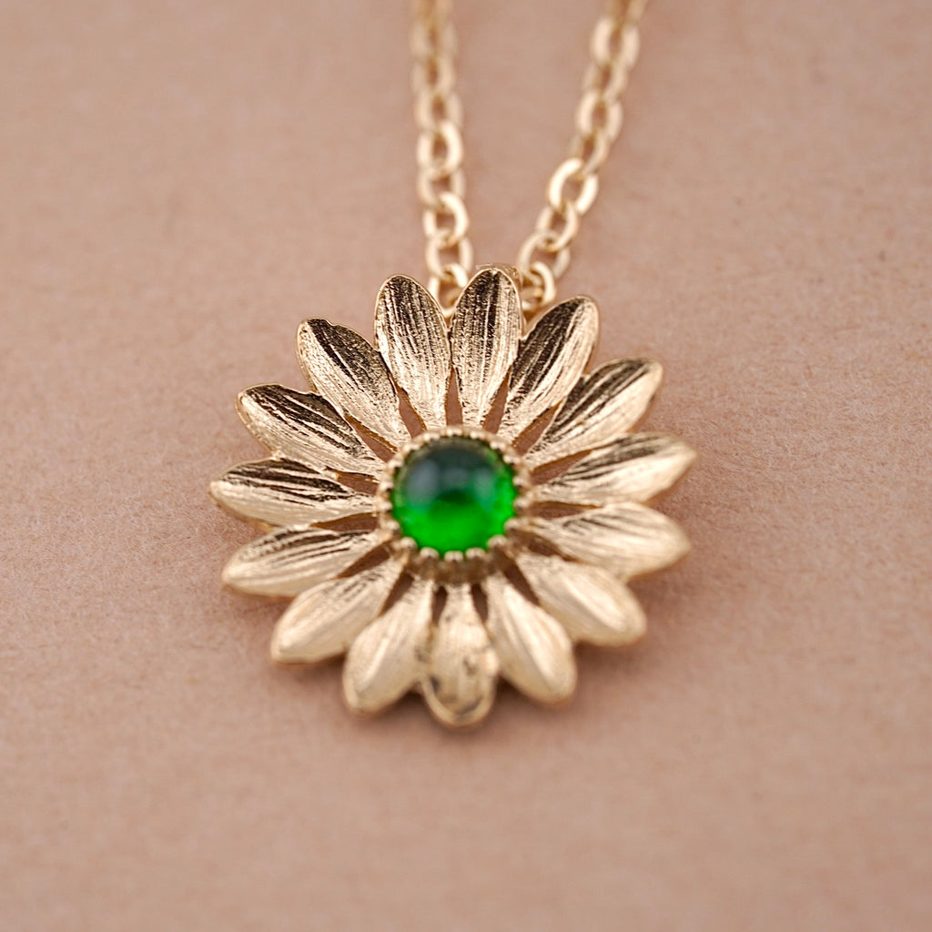 A close-up image of a 14K gold sunflower necklace with a vibrant green stone center, displayed against a clean, plain background. The delicate petals and polished gold finish highlight its elegant craftsmanship and symbolic beauty.
