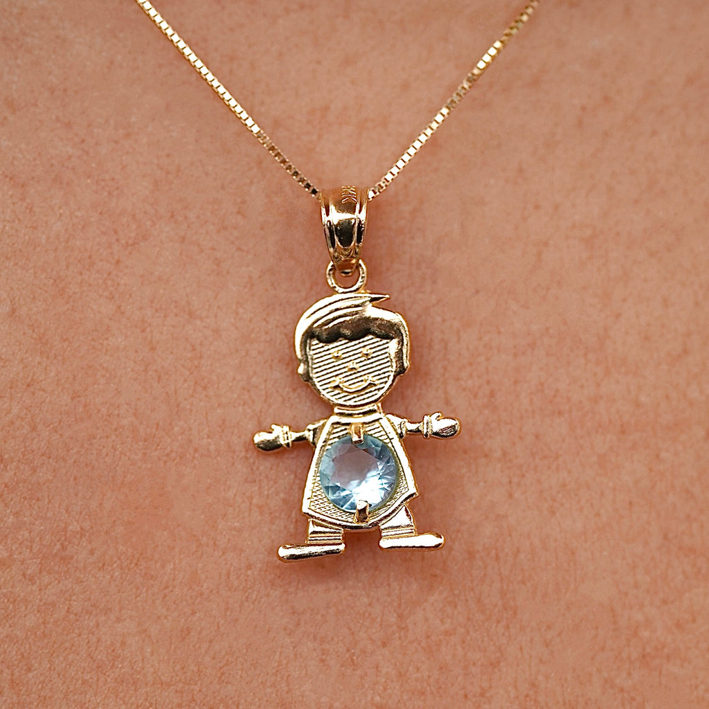 Model wearing a 14K solid gold boy charm necklace featuring a light blue cubic zirconia birthstone for March, layered with other gold chains, creating a stylish, personalized jewelry stack against a light sweater.