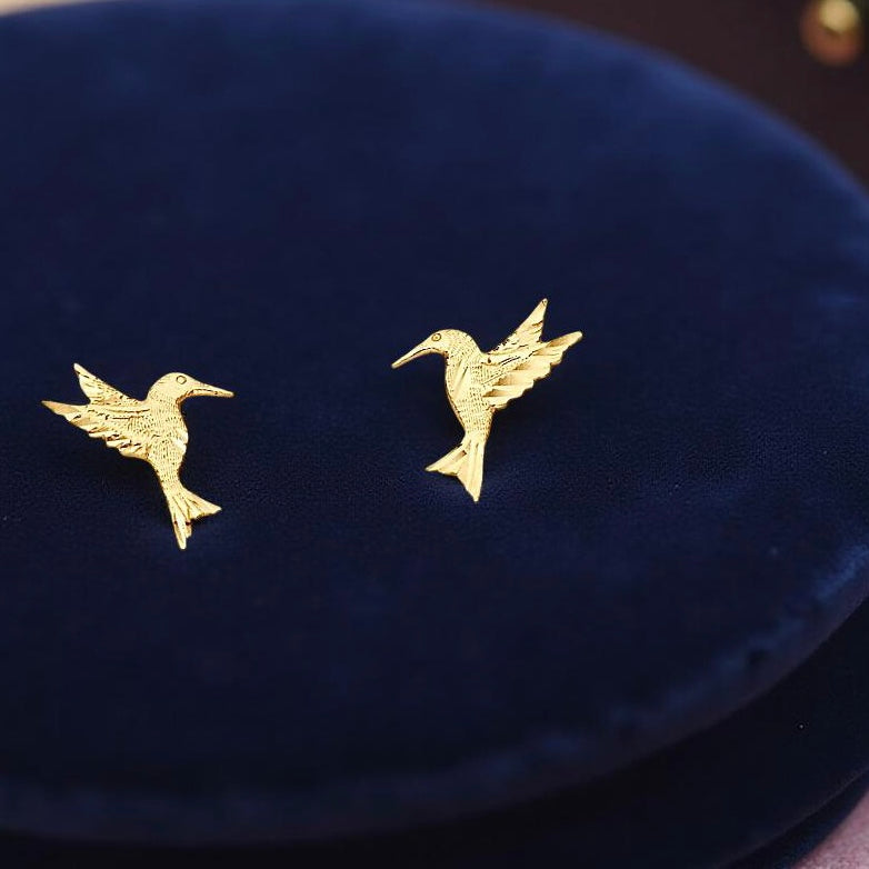 14K Gold Hummingbird Stud Earrings resting on dark blue velvet cloth, highlighting their intricate design and the warm gold tones of these elegant earrings.