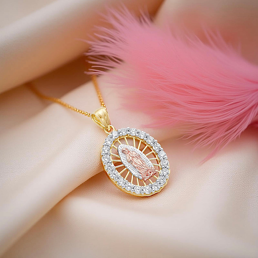 14K Gold Virgin of Guadalupe charm delicately placed on a soft pink textured cloth, highlighting the intricate details of the design and the brilliance of the surrounding cubic zirconia stones. The elegant fabric background enhances the warm tone of the yellow gold, creating a serene and graceful presentation of this spiritual piece.