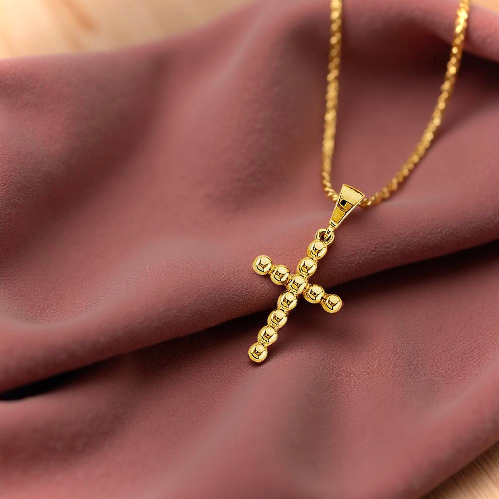 Close-up of a small beaded cross pendant in 14K yellow gold on dusty-rose fabric, showcasing its unique circular bead construction.