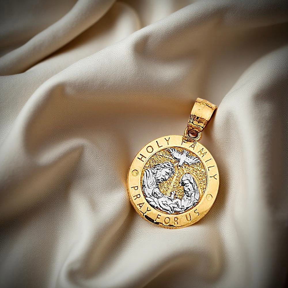 14K two-tone gold Holy Family pendant resting on neutral fabric, highlighting the intricate engraved artwork and radiant finish in natural lighting.