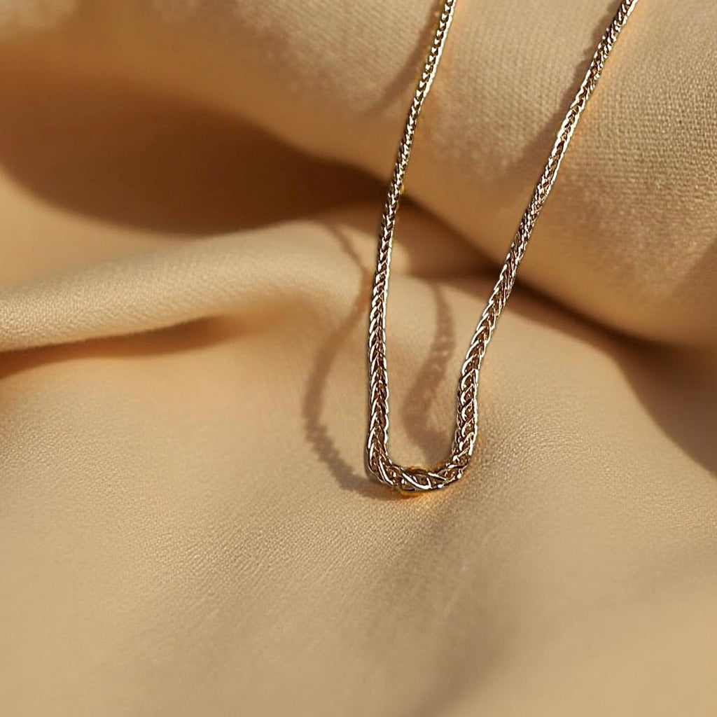 Delicate 14K yellow gold wheat chain with diamond-cut shine, laid gracefully on a textured off-beige fabric background for a soft, elegant presentation.
