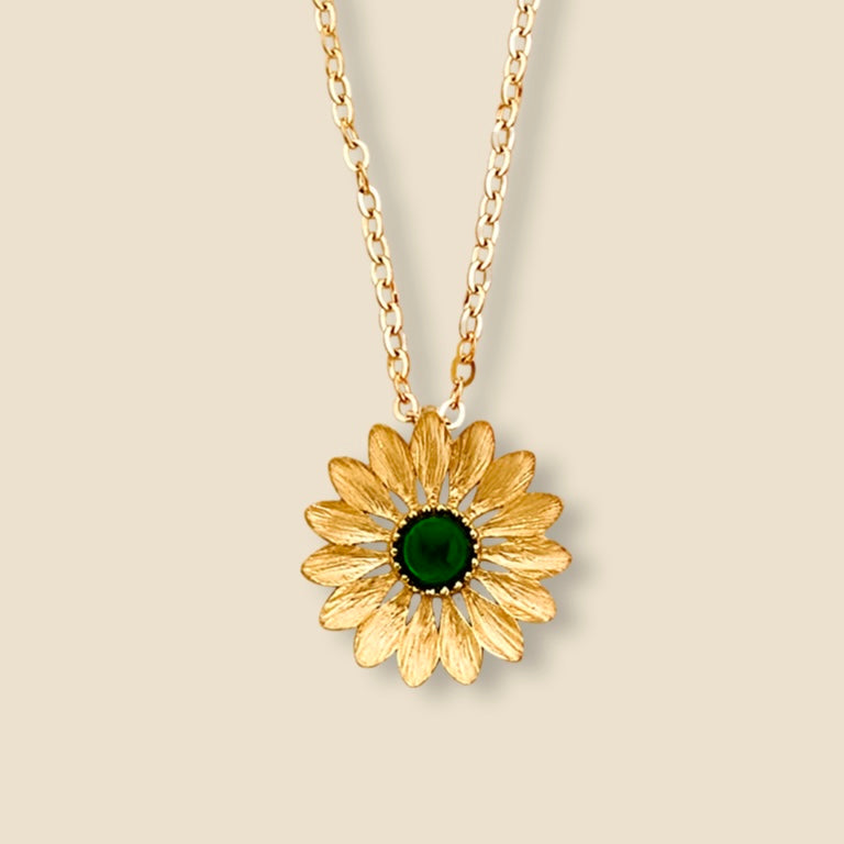 A close-up image of a 14K gold sunflower necklace with a vibrant green stone center, displayed against a clean, plain background. The delicate petals and polished gold finish highlight its elegant craftsmanship and symbolic beauty.