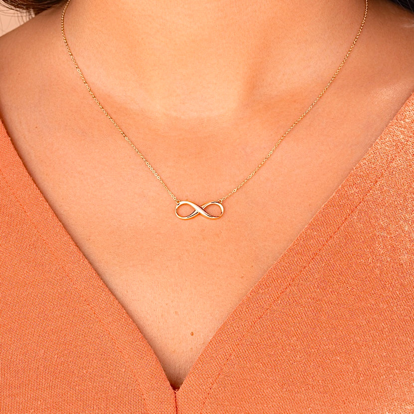 A woman wearing a 14K solid gold infinity necklace, the delicate gold chain resting just below her collarbone. The infinity symbol adds a touch of minimalist elegance, making it perfect for everyday wear or special occasions.