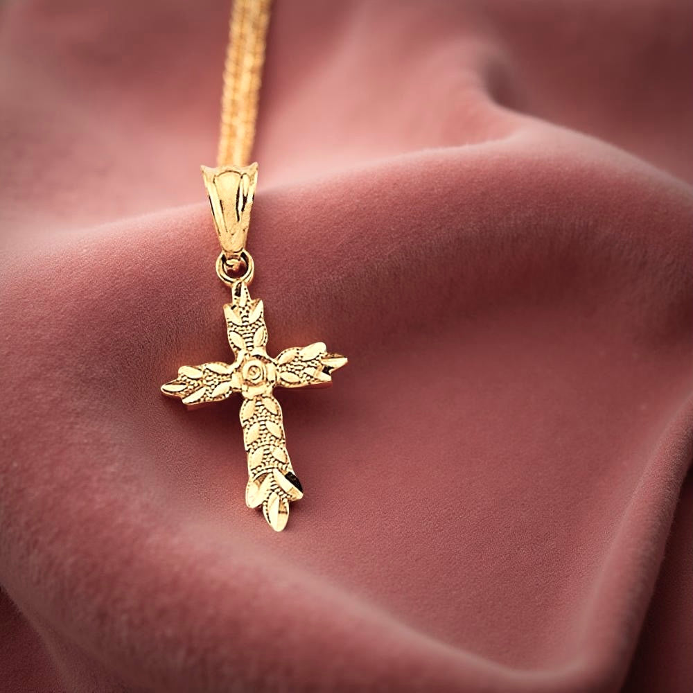 14K yellow gold cross pendant featuring delicate leaf accents and a rose flower at the center, resting on an dusty-rose textured cloth with soft shadows highlighting its botanical details.