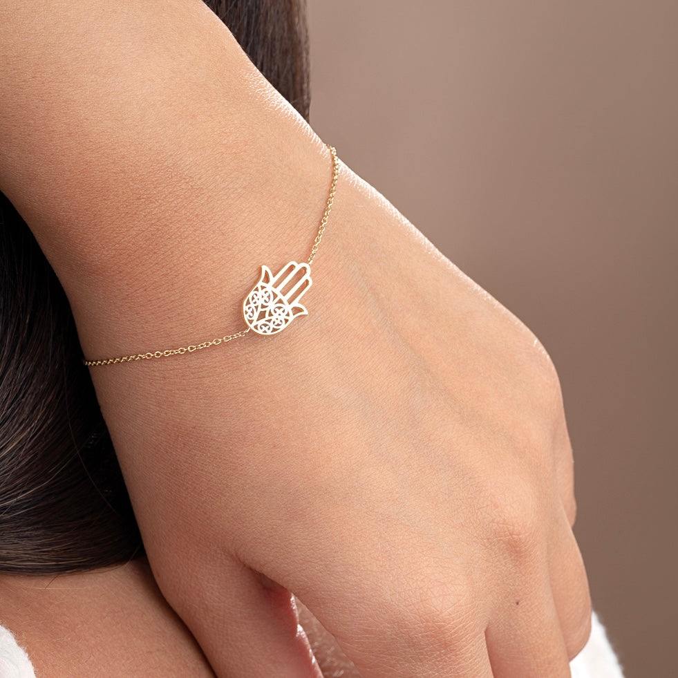 Model wearing a delicate 14K yellow gold Hamsa hand bracelet on wrist, showcasing its dainty charm and spiritual elegance against natural skin tone.