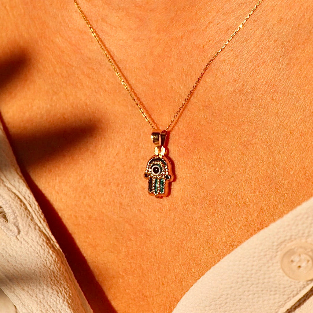 A model showcases a 14K yellow gold Hamsa pendant with a radiant blue Evil Eye centered in the palm. The delicate charm sits elegantly on the chest, adding a meaningful and stylish accent to any outfit.