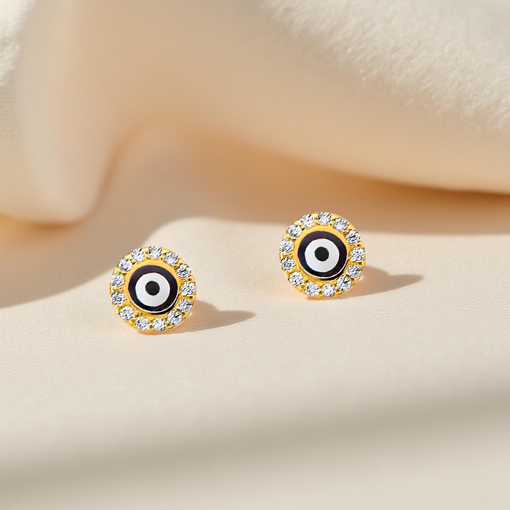 A pair of dainty 14K yellow gold evil eye stud earrings, featuring a brilliant cubic zirconia center, carefully positioned on soft off-white cloth with subtle fabric texture highlighting their delicate craftsmanship and warm gold finish.
