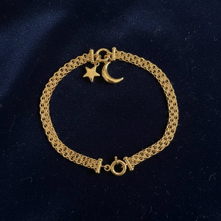 A 14K solid yellow gold star and moon bracelet laid on dark blue velvet cloth, highlighting the shine of the celestial charms against the dark fabric.