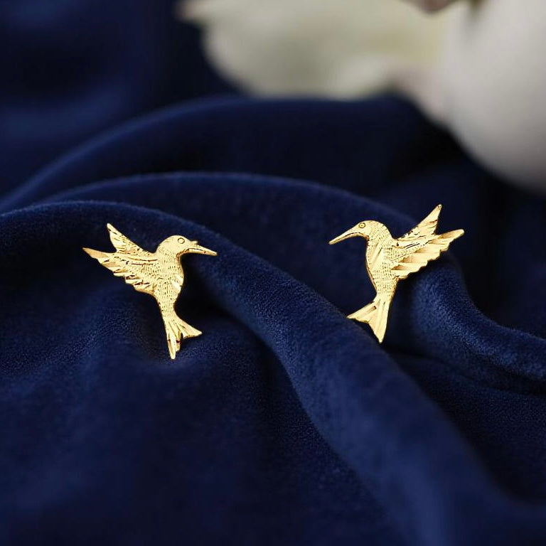 14K Gold Hummingbird Stud Earrings resting on dark blue velvet cloth, highlighting their intricate design and the warm gold tones of these elegant earrings.