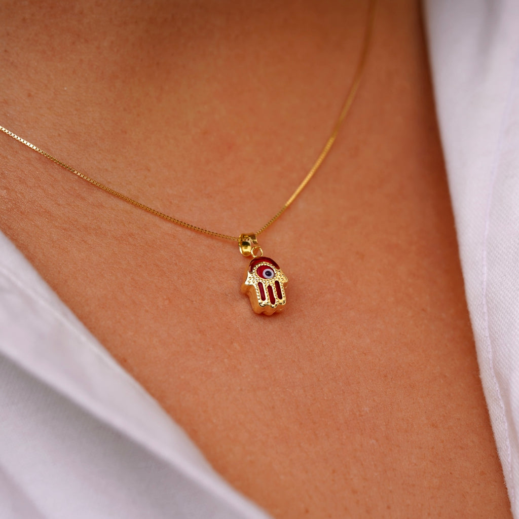 A model wears a 14K gold Hamsa pendant featuring a bold red Evil Eye, resting gracefully on the neckline. The vibrant red detail adds a unique touch of energy and charm to the protective talisman.
