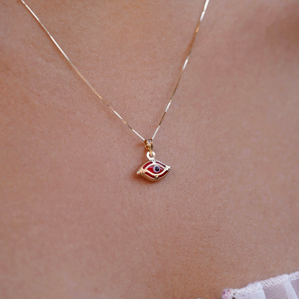 A model wearing a delicate 14K yellow gold red evil eye pendant featuring a brilliant red cubic zirconia center, resting elegantly on her neckline. The fine gold chain and polished finish reflect soft light, highlighting the pendant’s symbolic protection and timeless charm.