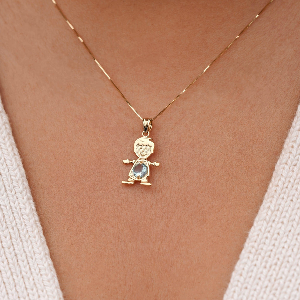 Model wearing a 14K solid gold boy charm necklace featuring a light blue cubic zirconia birthstone for March, layered with other gold chains, creating a stylish, personalized jewelry stack against a light sweater.