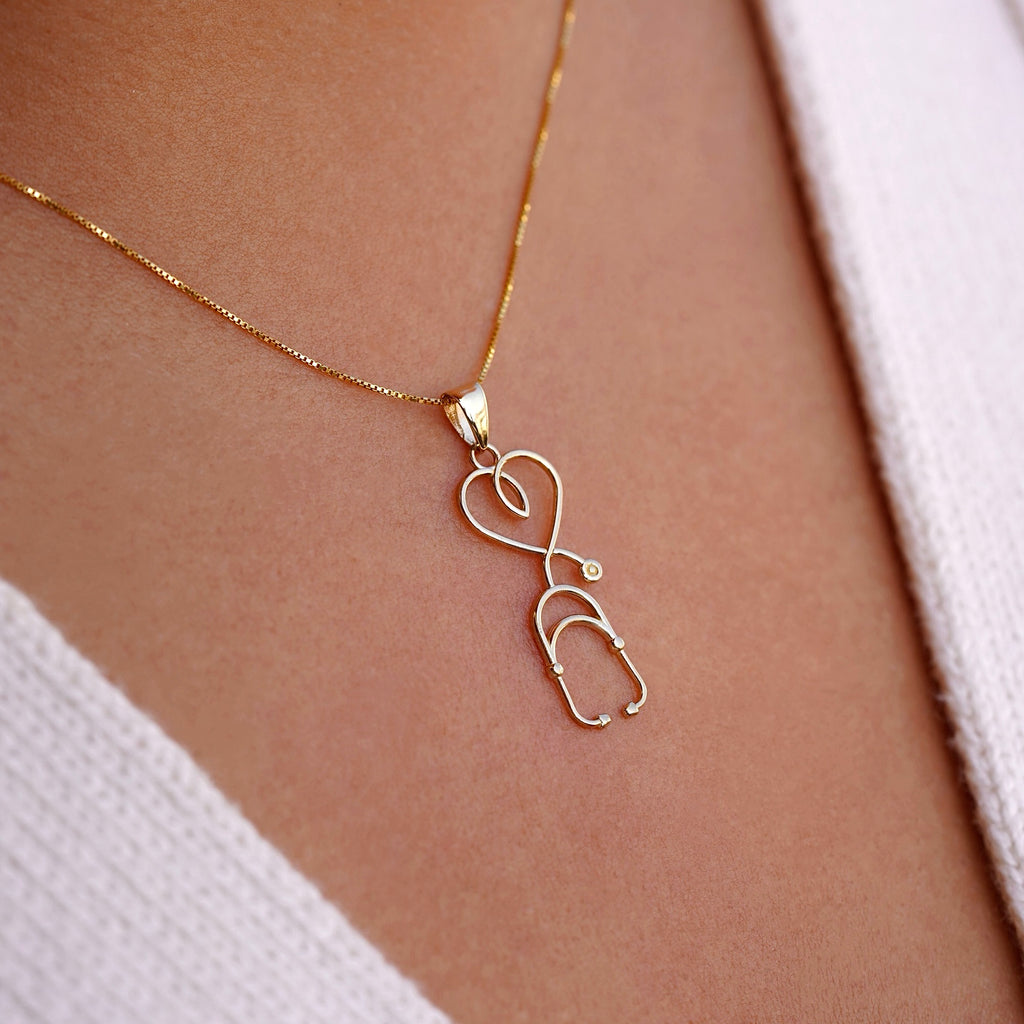 A model wears the 14K yellow gold stethoscope pendant on a delicate gold chain, showcasing its sleek vertical design against a neutral outfit.