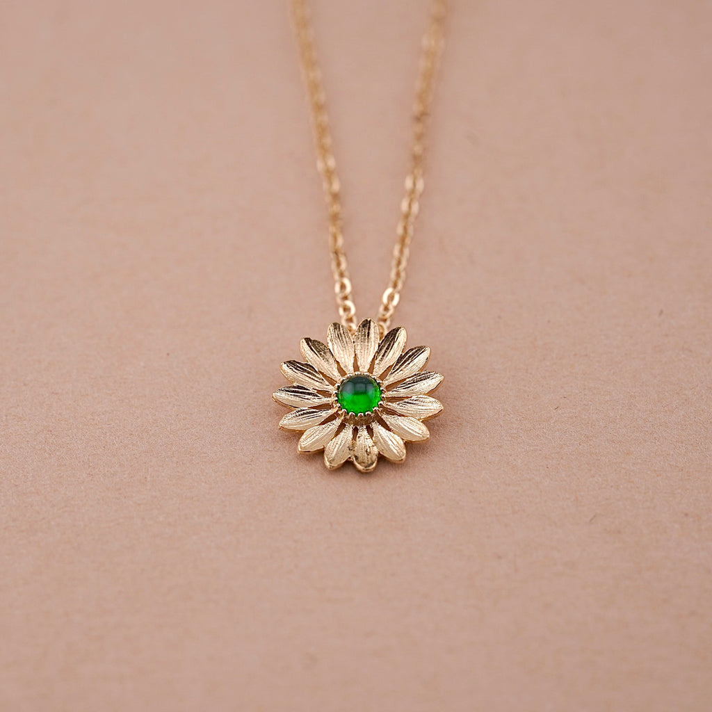 A close-up image of a 14K gold sunflower necklace with a vibrant green stone center, displayed against a clean, plain background. The delicate petals and polished gold finish highlight its elegant craftsmanship and symbolic beauty.