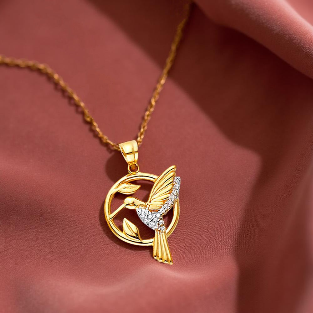 Close-up of 14K gold hummingbird pendant resting on dusty rose fabric, highlighting its graceful shape and polished shine.