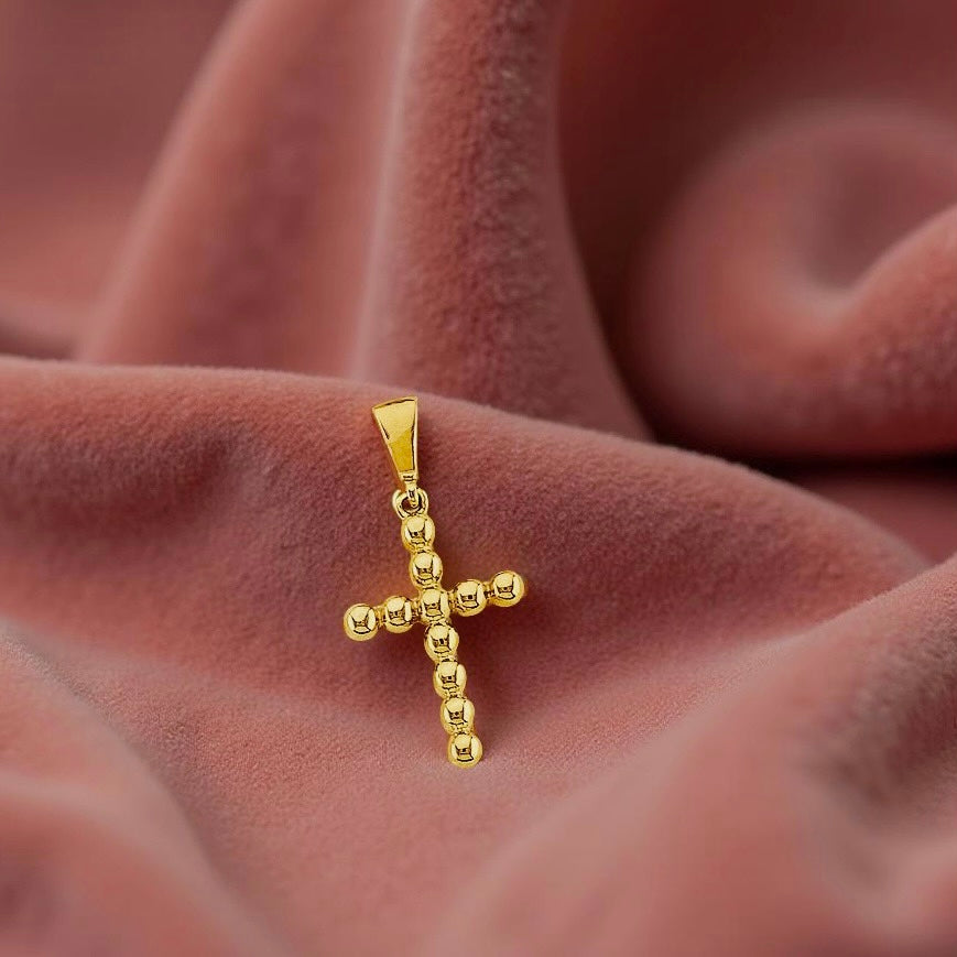 Close-up of a small beaded cross pendant in 14K yellow gold on dusty-rose fabric, showcasing its unique circular bead construction.