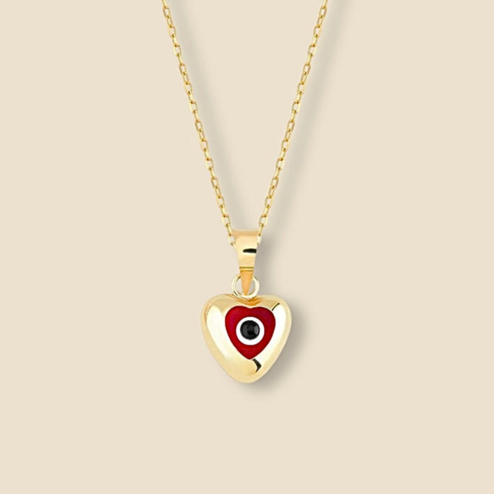 A close-up of the 14K yellow gold heart pendant with a deep red enamel eye center, resting on neutral and clean background, symbolizing classic protection and offering a minimalist yet bold pop of color.