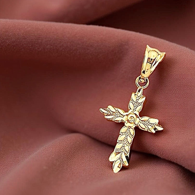 14K yellow gold cross pendant featuring delicate leaf accents and a rose flower at the center, resting on an dusty-rose textured cloth with soft shadows highlighting its botanical details.
