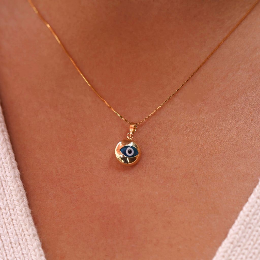 A model wearing a 14K yellow gold evil eye pendant with a blue zirconia center on a fine gold chain, resting elegantly at her collarbone. The polished gold finish reflects light beautifully, highlighting the pendant’s delicate craftsmanship, symbolic protection, and subtle luxury from Tehrani Jewelry.