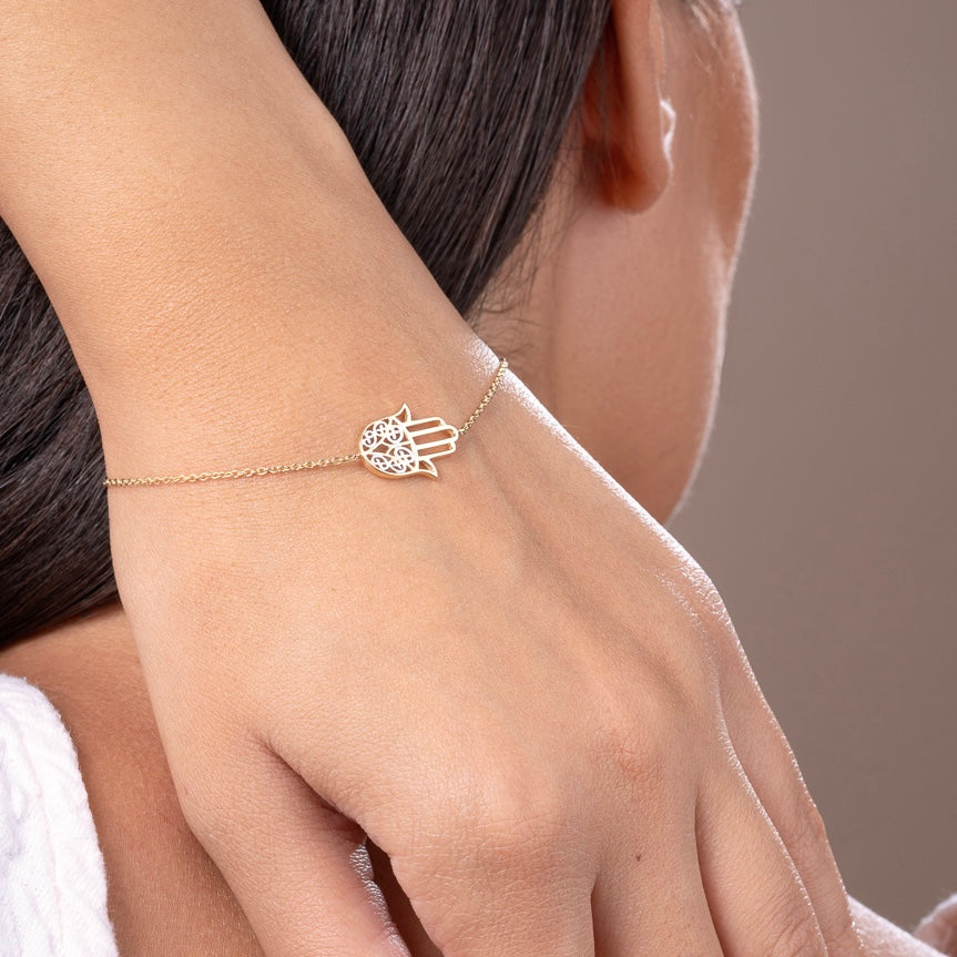 Model wearing a delicate 14K yellow gold Hamsa hand bracelet on wrist, showcasing its dainty charm and spiritual elegance against natural skin tone.