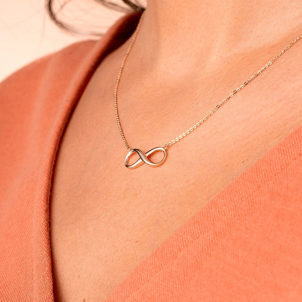 A woman wearing a 14K solid gold infinity necklace, the delicate gold chain resting just below her collarbone. The infinity symbol adds a touch of minimalist elegance, making it perfect for everyday wear or special occasions.