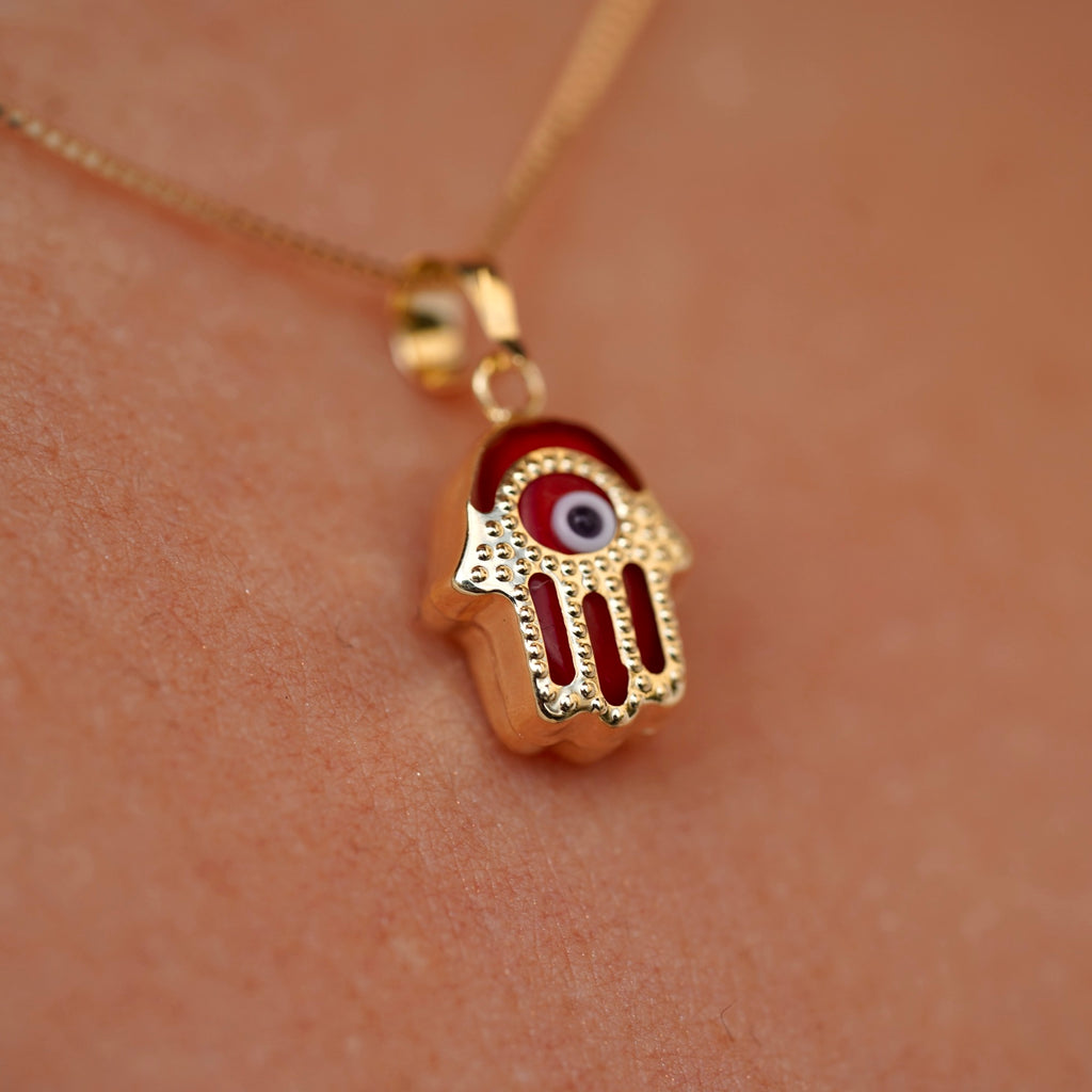 A model wears a 14K gold Hamsa pendant featuring a bold red Evil Eye, resting gracefully on the neckline. The vibrant red detail adds a unique touch of energy and charm to the protective talisman.