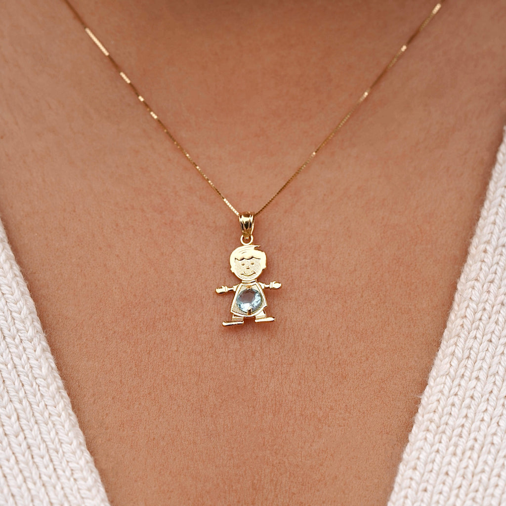Model wearing a 14K solid gold boy charm necklace featuring a light blue cubic zirconia birthstone for March, layered with other gold chains, creating a stylish, personalized jewelry stack against a light sweater.