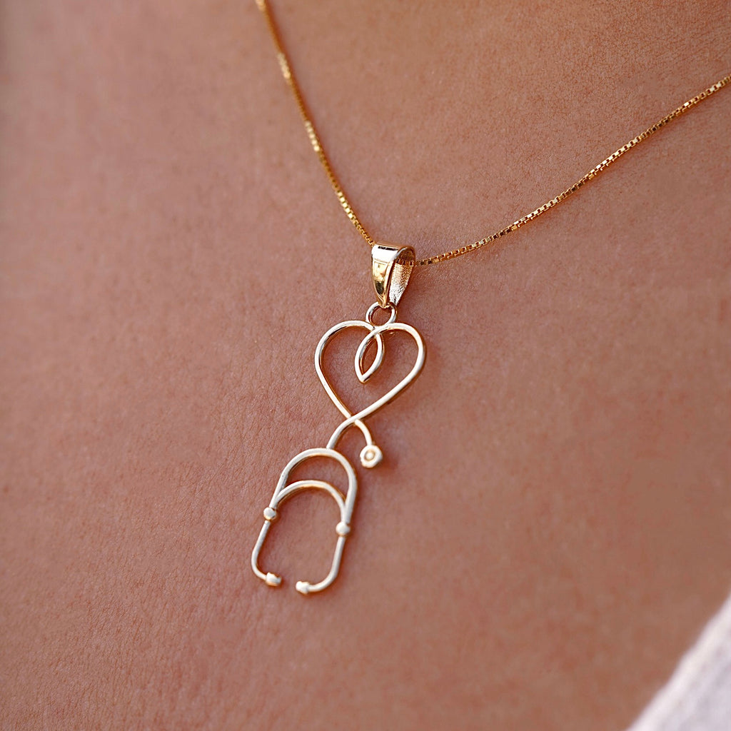 A model wears the 14K yellow gold stethoscope pendant on a delicate gold chain, showcasing its sleek vertical design against a neutral outfit.