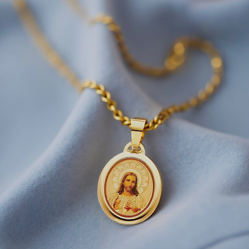 14K gold Sacred Heart of Jesus pendant displayed on soft blue fabric with red enamel details glowing in natural light.