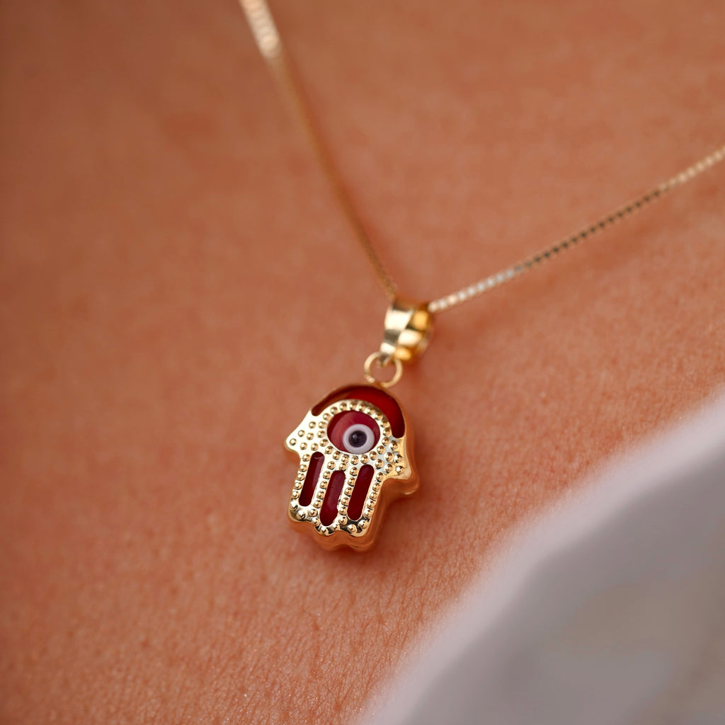 A model wears a 14K gold Hamsa pendant featuring a bold red Evil Eye, resting gracefully on the neckline. The vibrant red detail adds a unique touch of energy and charm to the protective talisman.