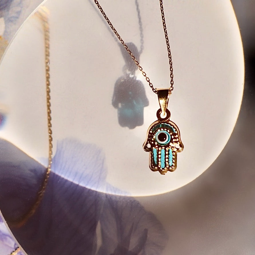 A 14K yellow gold Hamsa pendant with a striking blue Evil Eye at its center, displayed on a neutral backdrop. The polished gold and vivid blue eye create a powerful symbol of protection and style.