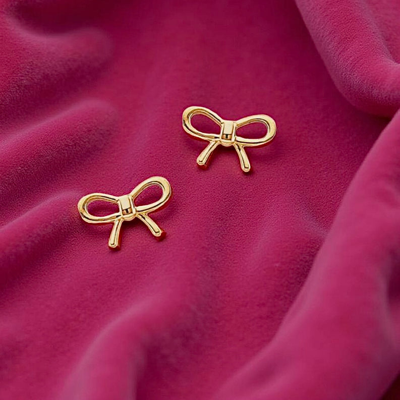 14K gold ribbon stud earrings delicately placed on magenta velvet cloth, showcasing their dainty bow shape and fine gold finish.
