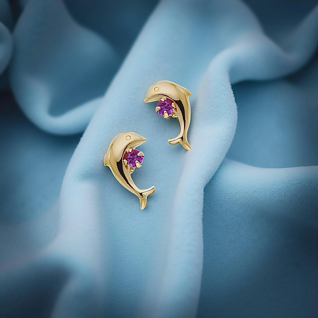 14K gold dolphin earrings with red CZ stones resting on a soft blue fabric, showcasing their ocean-inspired design and fine polish.