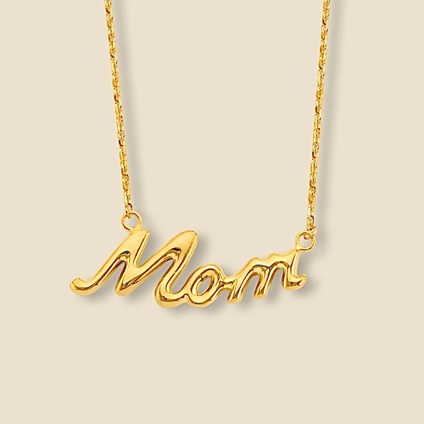 Close-up of 14K Yellow Gold “Mom” Necklace with a high-polish finish, laid flat on a plain surface to highlight the warm shine and smooth craftsmanship of the solid gold word pendant.