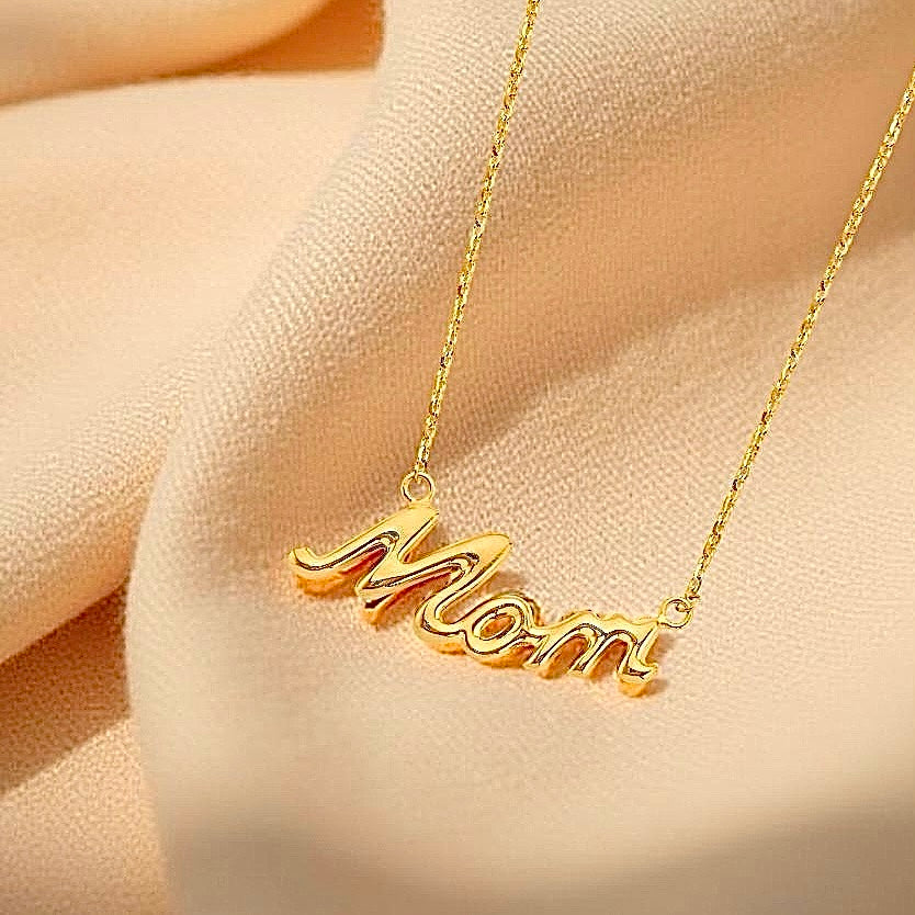 14K Yellow Gold “Mom” Necklace displayed on soft off-cream textured fabric, emphasizing the natural golden hue and graceful curves of the word pendant for a cozy, gift-ready presentation.