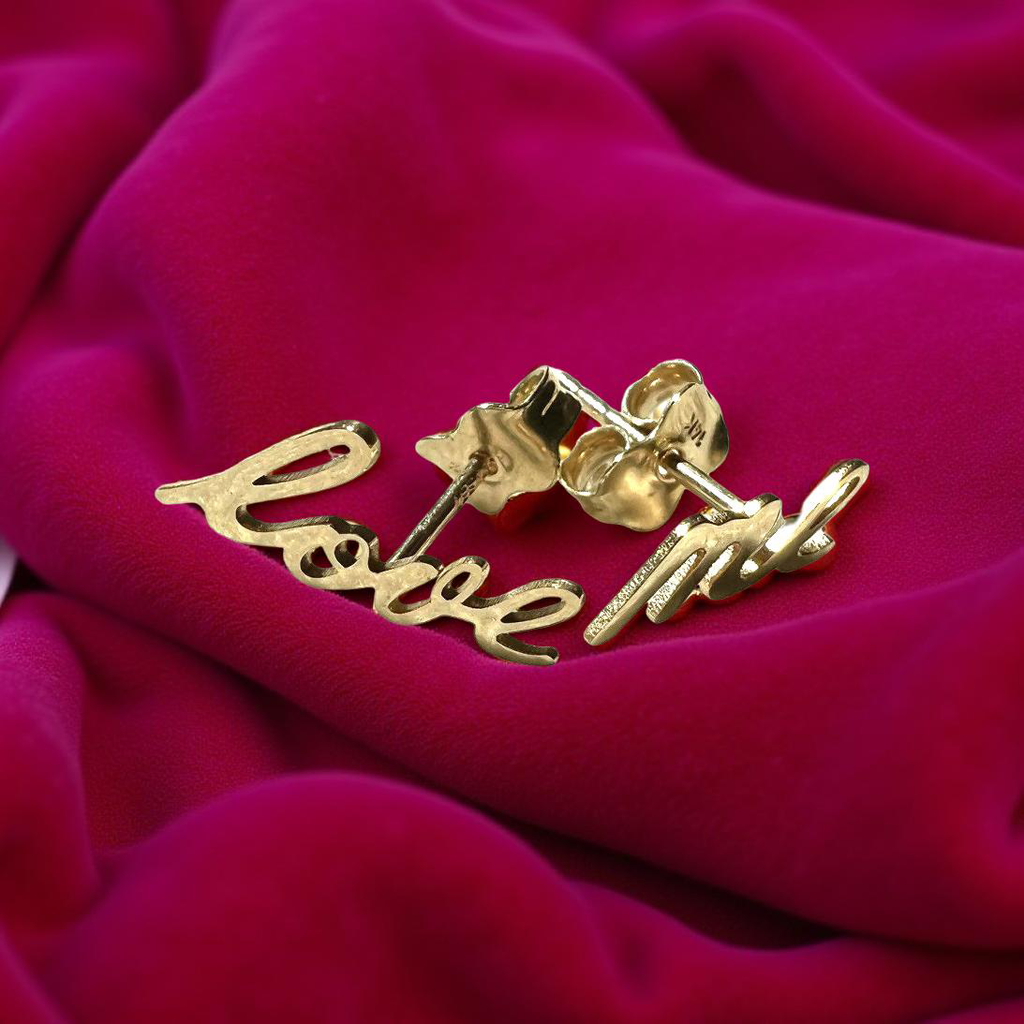 lose-up of two “Love Me” 14K yellow-gold script earrings resting on a magenta velvet fabric, highlighting their polished shine and delicate lettering.