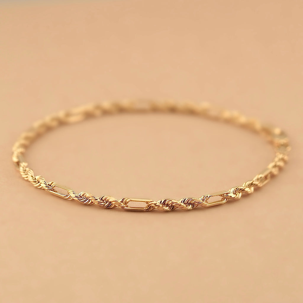 Solid 14K yellow gold Figaro rope bracelet shown on a beige background, focusing on its interwoven chain and rich golden tone.