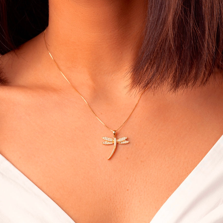 A close-up of a model wearing the 14K gold dragonfly pendant on a fine gold chain. The pendant sits gracefully on the collarbone, catching the light with its cubic zirconia details for a sophisticated, nature-inspired look.