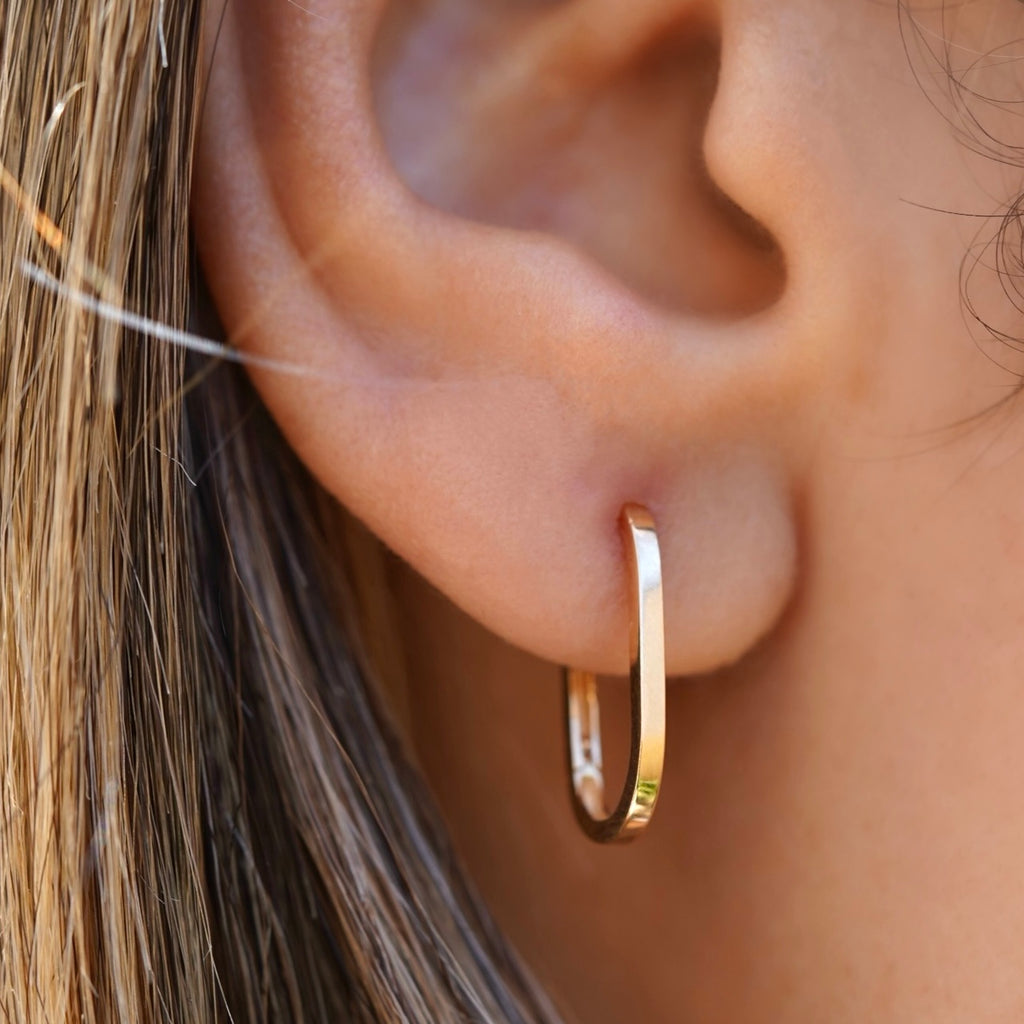 A model wearing 14K Yellow Gold Shiny Paperclip Huggies Earrings. The sleek and minimalist paperclip-inspired design is highlighted as they rest elegantly on the model’s ears, adding a chic and modern touch to their look.