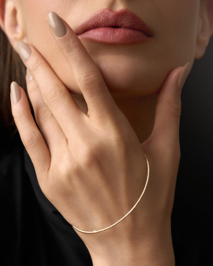 A model wearing 14K gold braided bangle on her wrist, showcasing its delicate 1.5mm rope-inspired design. The bangle shines in a polished yellow gold finish, highlighting its elegant, lightweight, and dainty texture. The image demonstrates how the bangle can be worn alone for a minimalist look or stacked with other bracelets for a layered, sophisticated style, perfect for everyday wear or special occasions.