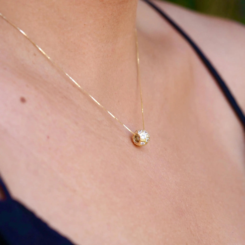 A woman elegantly wears the 14K Gold Bead Charm Pendant on a delicate gold chain. The diamond-cut finish and cubic zirconia sparkle as they catch the light, adding a touch of timeless sophistication to her outfit. The pendant rests gracefully on her collarbone, showcasing its minimalist yet luxurious design.